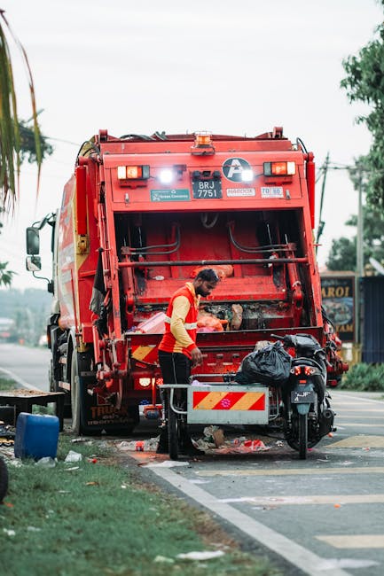 A sanitation worker in a red and yellow uniform is seen operating a large red refuse collection vehicle parked on the side of a street during daytime. The worker is standing close to the open rear com